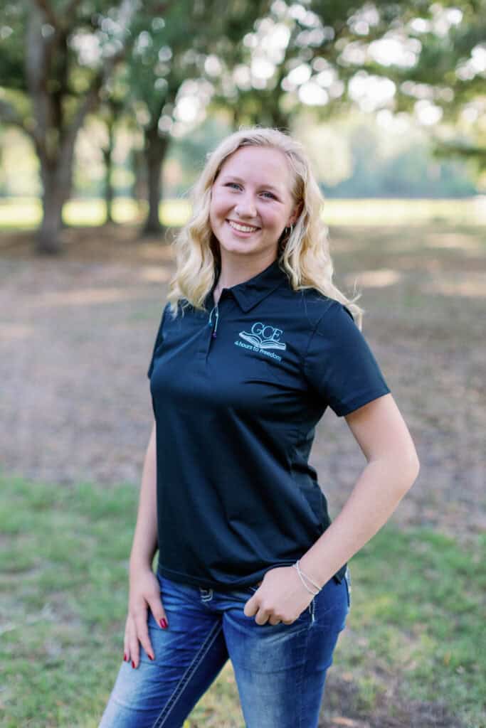 A young woman smiling outdoors, wearing a GCE polo shirt, representing a student in a classical homeschool program.