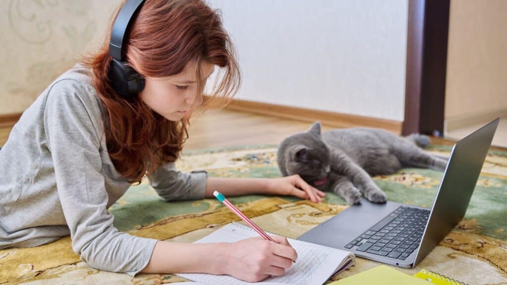 A young girl with headphones studies online with her cat nearby, illustrating a hybrid homeschool environment.