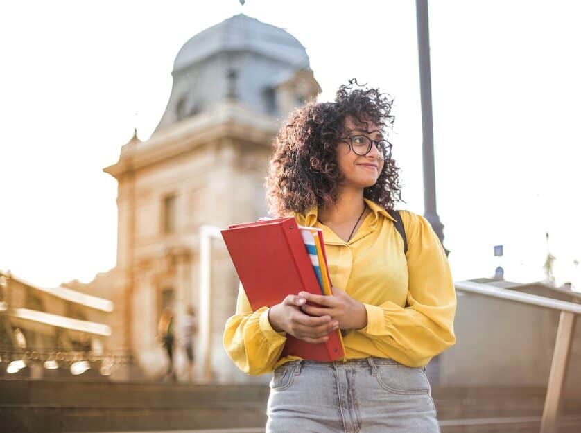 A young girl with curly hair holding textbooks, standing outside a historic building, representing classical education stages.