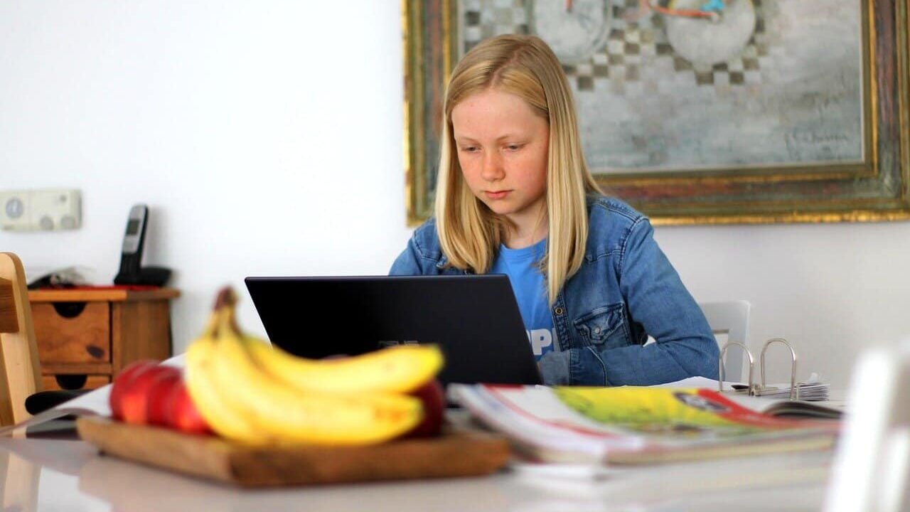 A young girl studying on a laptop at home, surrounded by books and school supplies, in a classical education setting.