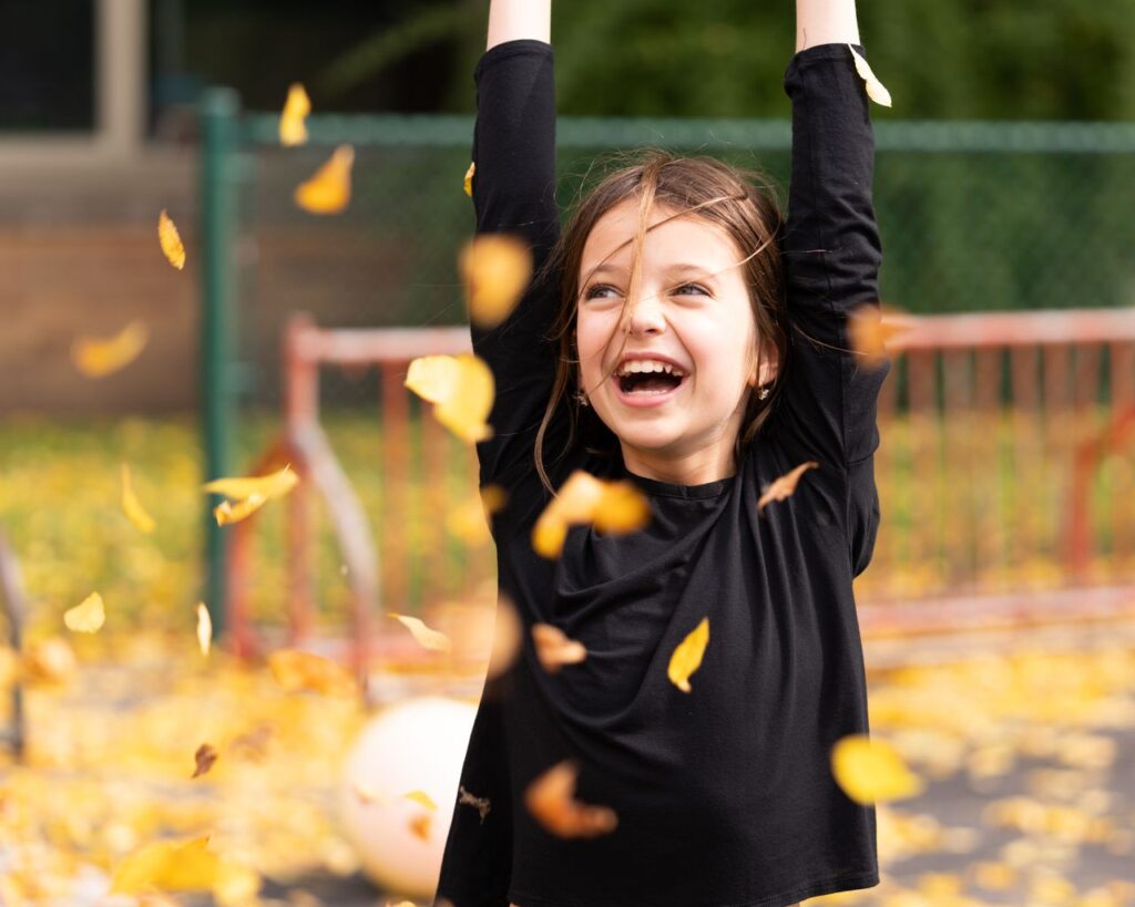 pexels-photo-4680787-4680787 Happy smiling girl with shaggy brown hair in black shirt playing with autumn foliage at daytime